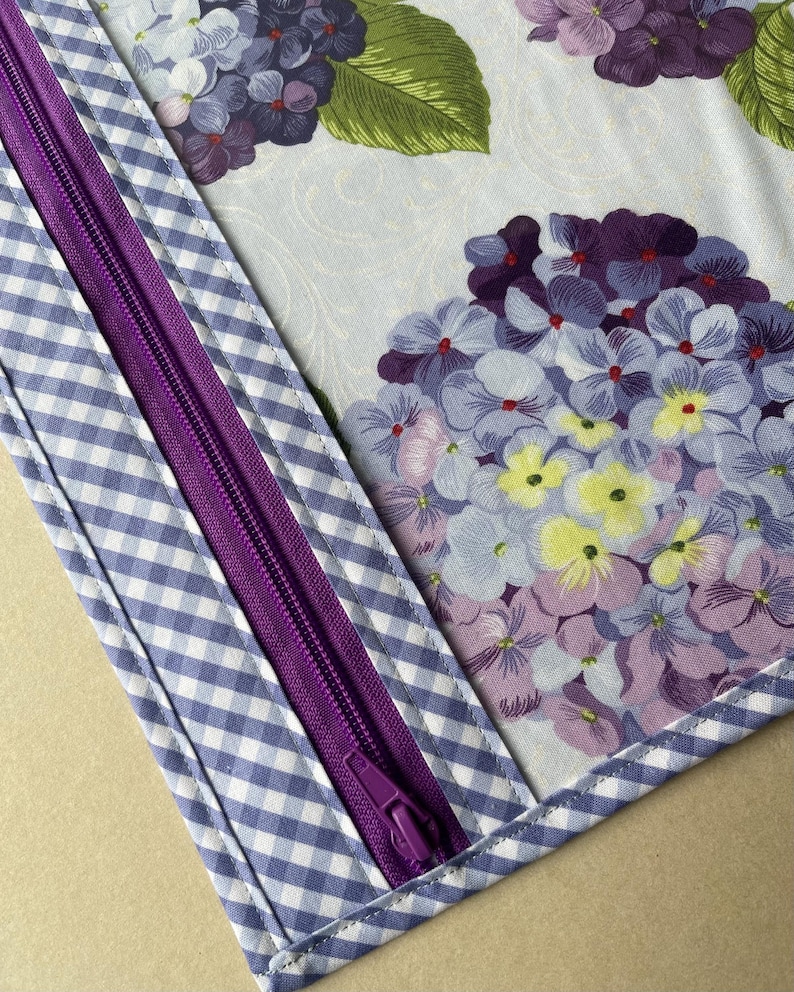 May include: A close-up of a fabric item featuring a purple zipper and a blue and white gingham pattern. The main fabric displays a floral design with purple, blue, and yellow hydrangea blooms and green leaves.