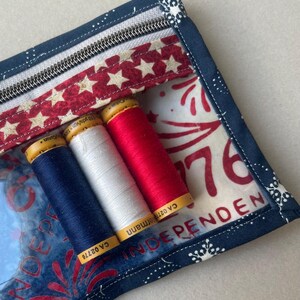 May include: A patriotic-themed sewing kit with a clear front, featuring spools of blue, white, and red thread. The kit has a zipper closure and is decorated with stars and the word "INDEPENDENCE". The background has a firework design.