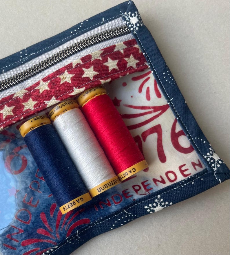May include: A patriotic-themed sewing kit with a clear front, featuring spools of blue, white, and red thread. The kit has a zipper closure and is decorated with stars and the word "INDEPENDENCE". The background has a firework design.