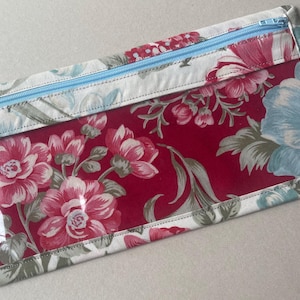 May include: A rectangular floral print pouch with a clear window. The pouch features a red background with pink and blue flowers and green leaves. The edges are trimmed with a floral fabric and a light blue zipper.