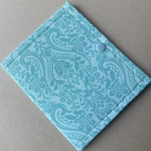 May include: A square fabric pouch in a light turquoise blue color featuring a tonal damask floral pattern. The pouch is constructed with visible stitched edges and includes a single matching light turquoise blue plastic snap button closure.