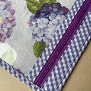 May include: Close-up of a fabric item featuring a floral pattern of purple and white hydrangeas on a light background. A purple zipper is sewn into a section of blue and white gingham fabric.