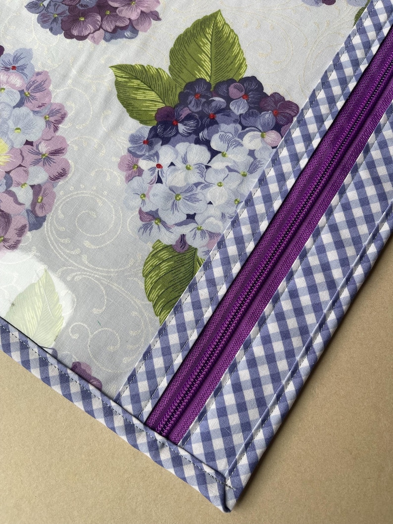May include: Close-up of a fabric item featuring a floral pattern of purple and white hydrangeas on a light background. A purple zipper is sewn into a section of blue and white gingham fabric.
