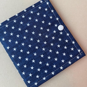 May include: A square fabric pouch featuring a dark blue background with a repeating pattern of small white stars. The pouch is constructed with visible stitching along the edges and is secured with a single white plastic snap button closure.