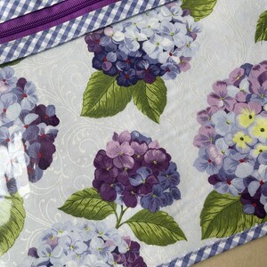 May include: A floral pattern featuring large hydrangea blooms in shades of purple, lavender, and white, with green leaves. The design is on a light blue background with a subtle swirl pattern. A purple zipper and blue and white gingham trim complete the design.