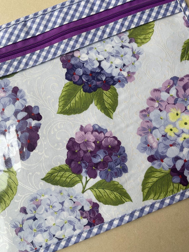 May include: A floral pattern featuring large hydrangea blooms in shades of purple, lavender, and white, with green leaves. The design is on a light blue background with a subtle swirl pattern. A purple zipper and blue and white gingham trim complete the design.