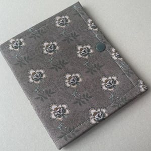 May include: A gray fabric journal cover with a floral pattern. The cover features a repeating pattern of white and brown flowers with green leaves and stems. A green button secures the cover closed. The edges are neatly stitched.
