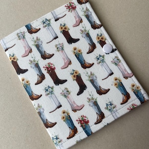 May include: A rectangular fabric cover featuring a repeating pattern of cowboy boots in various colors, each filled with floral arrangements of sunflowers and wildflowers. The cover is off-white and includes a single white snap button closure.