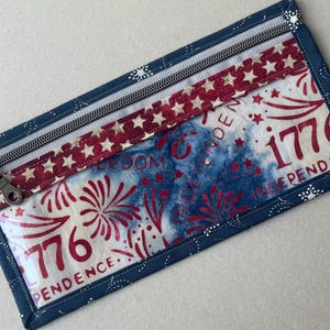 May include: Rectangular zippered pouch with a patriotic theme. The pouch features a blue border with white star accents, a red and white star-patterned band, and a central panel with red fireworks and the text "1776 INDEPENDENCE".