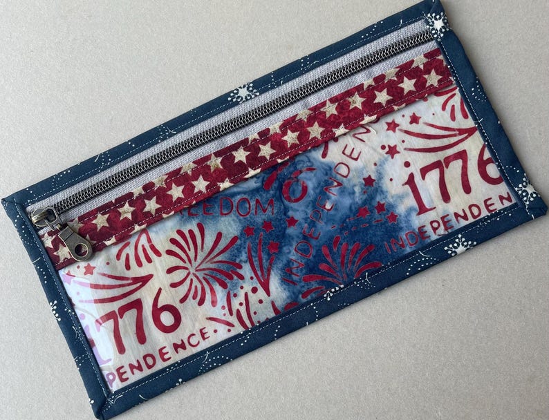 May include: Rectangular zippered pouch with a patriotic theme. The pouch features a blue border with white star accents, a red and white star-patterned band, and a central panel with red fireworks and the text "1776 INDEPENDENCE".