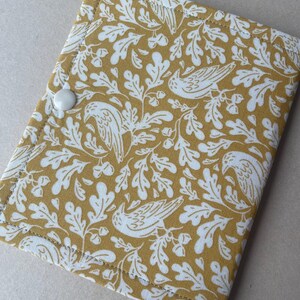 May include: A rectangular fabric case featuring a mustard yellow background with a white repeating pattern of birds, oak leaves, and acorns. The case is secured with a single white plastic snap button and has visible stitching along the edges.