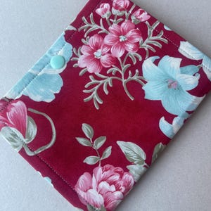 May include: A floral fabric item with a deep red background and a pattern of pink and light blue flowers. The item is secured with a light blue button. The edges are neatly stitched, suggesting a handmade or crafted product.