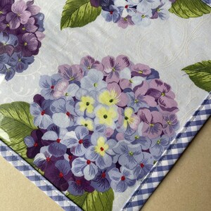 May include: A floral pattern featuring purple, lavender, and yellow hydrangea blooms with green leaves on a light blue background. The fabric is trimmed with a blue and white gingham border. The design is likely for home decor or a craft project.