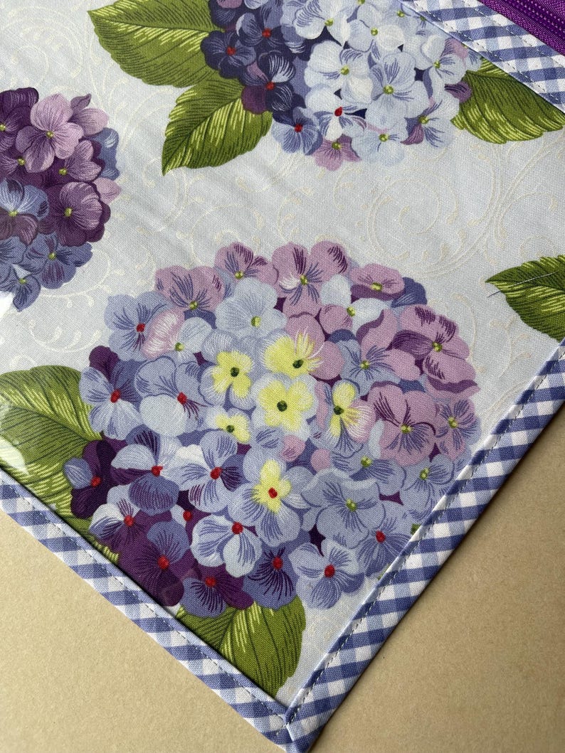 May include: A floral pattern featuring purple, lavender, and yellow hydrangea blooms with green leaves on a light blue background. The fabric is trimmed with a blue and white gingham border. The design is likely for home decor or a craft project.