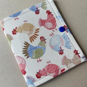 May include: A fabric item with a repeating pattern of colorful rooster illustrations in red, blue, green, and tan on a white background. The item is rectangular with a blue button closure and stitched edges. The rooster design gives a farmhouse or country theme.