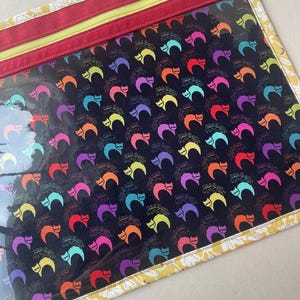 May include: A rectangular zippered pouch featuring a clear vinyl front panel with a repeating pattern of colorful cats in orange, yellow, purple, pink, and teal on a black background. The pouch has a red fabric zipper border and yellow trim.