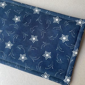 May include: A navy blue fabric item with a white snowflake and dotted pattern. The fabric is rectangular with a white stitched border. The design features stylized snowflakes and curved dotted lines, creating a wintery theme.