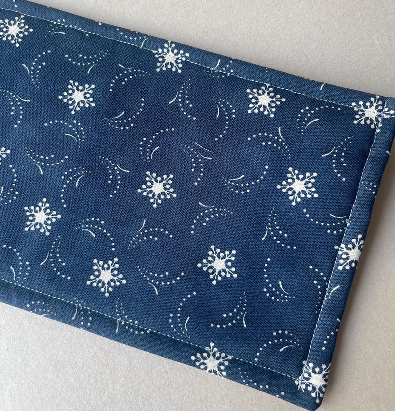 May include: A navy blue fabric item with a white snowflake and dotted pattern. The fabric is rectangular with a white stitched border. The design features stylized snowflakes and curved dotted lines, creating a wintery theme.