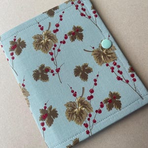 May include: A rectangular fabric cover in light blue featuring a repeating pattern of brown leaves and small red berries. The cover is secured with a single, solid light mint green plastic snap button located on the right side near the top edge.