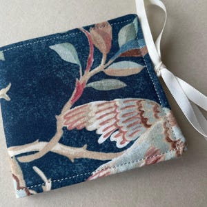 May include: A square fabric pouch featuring a dark blue background with a printed pattern of light brown branches, green leaves, and a bird with tan and white feathers. The pouch is finished with white stitching and a white satin ribbon tie.