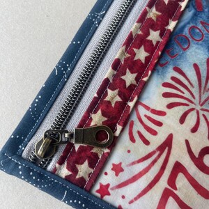 May include: Close-up of a patriotic-themed zippered pouch. The pouch features a navy blue fabric with white polka dots, a red and white star-patterned strip, and a white and blue fabric with red firework designs. The zipper is silver with a bronze pull.