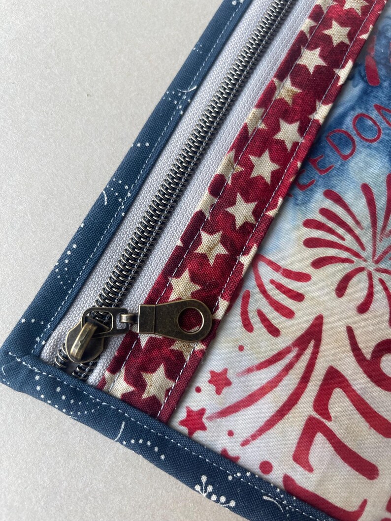 May include: Close-up of a patriotic-themed zippered pouch. The pouch features a navy blue fabric with white polka dots, a red and white star-patterned strip, and a white and blue fabric with red firework designs. The zipper is silver with a bronze pull.