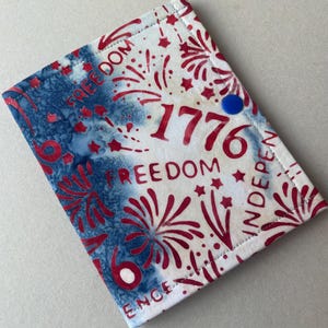 May include: A rectangular fabric item with a patriotic design. The design features red fireworks, stars, and the number "1776" on a white background. The left side has a blue and white tie-dye pattern. The word "FREEDOM" is repeated. A blue button is on the top right.