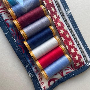 May include: A fabric sewing kit with a zipper, containing spools of thread in various colors: red, white, blue, and gray. The kit has a patriotic theme with a red and white star pattern and blue fabric with white floral designs. The word "INDEPENDENCE" is visible.