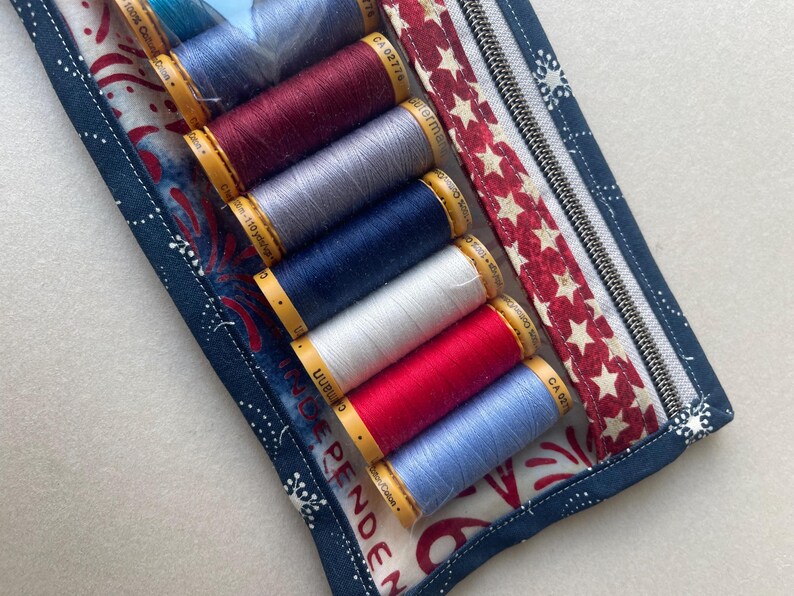 May include: A fabric sewing kit with a zipper, containing spools of thread in various colors: red, white, blue, and gray. The kit has a patriotic theme with a red and white star pattern and blue fabric with white floral designs. The word "INDEPENDENCE" is visible.