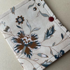May include: A beige fabric item with a floral pattern in brown, teal, and gray. The design features large flowers and leaves. A small, round, red button is on the corner. The item is likely a small case or cover.