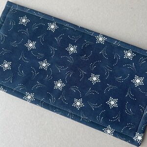 May include: A navy blue fabric placemat with a white snowflake and dotted design. The placemat is rectangular with a stitched border. The pattern features white snowflakes and dotted lines on a dark blue background.