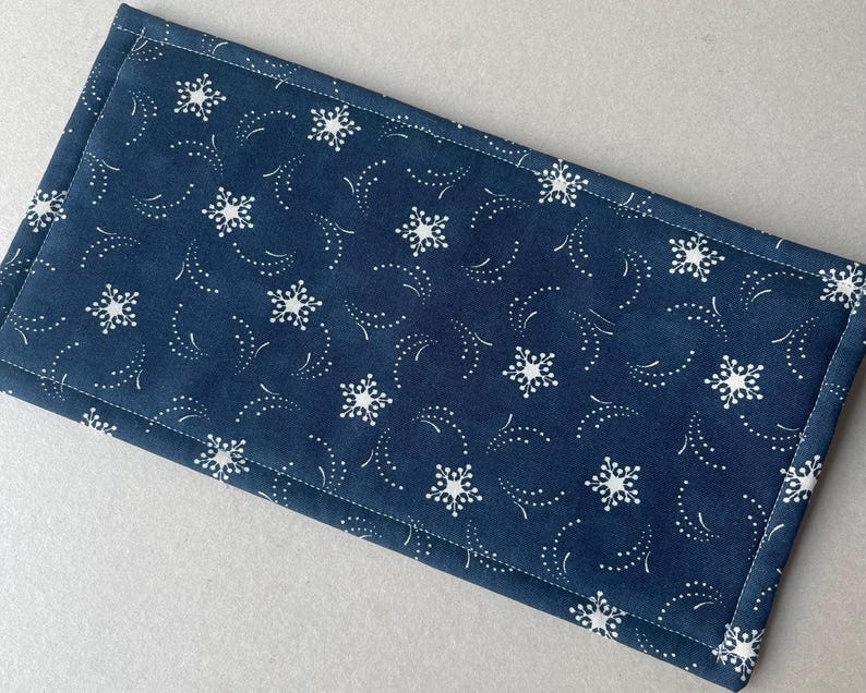 May include: A navy blue fabric placemat with a white snowflake and dotted design. The placemat is rectangular with a stitched border. The pattern features white snowflakes and dotted lines on a dark blue background.