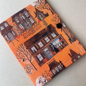 May include: A rectangular fabric cover featuring an orange background with a repeating pattern of brown houses, bare trees, pumpkins, and silhouettes of figures in hats. The cover is secured with a single black circular snap button closure.