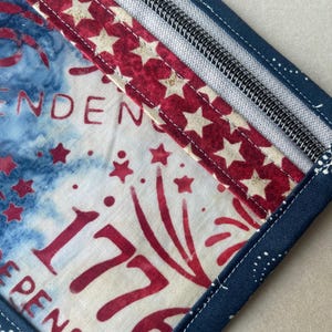 May include: A patriotic, square-shaped fabric item with a zipper, featuring a design of red stars on a white background, and the number "1776" in red. The item has a blue border with white polka dots and the word "INDEPENDENCE".