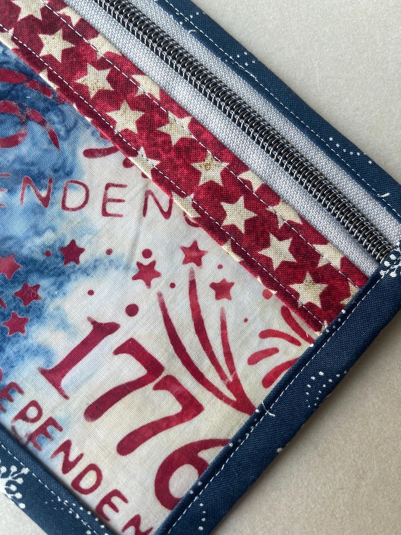 May include: A patriotic, square-shaped fabric item with a zipper, featuring a design of red stars on a white background, and the number "1776" in red. The item has a blue border with white polka dots and the word "INDEPENDENCE".