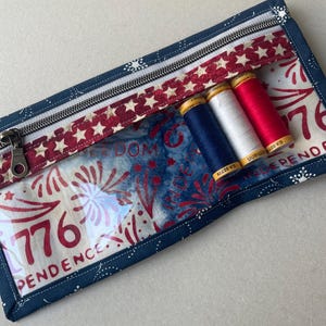 May include: A rectangular, patriotic-themed zippered pouch with a clear front panel. The pouch features a navy blue border, a red and white star-patterned accent, and a zipper. Inside are three spools of thread in red, white, and blue.