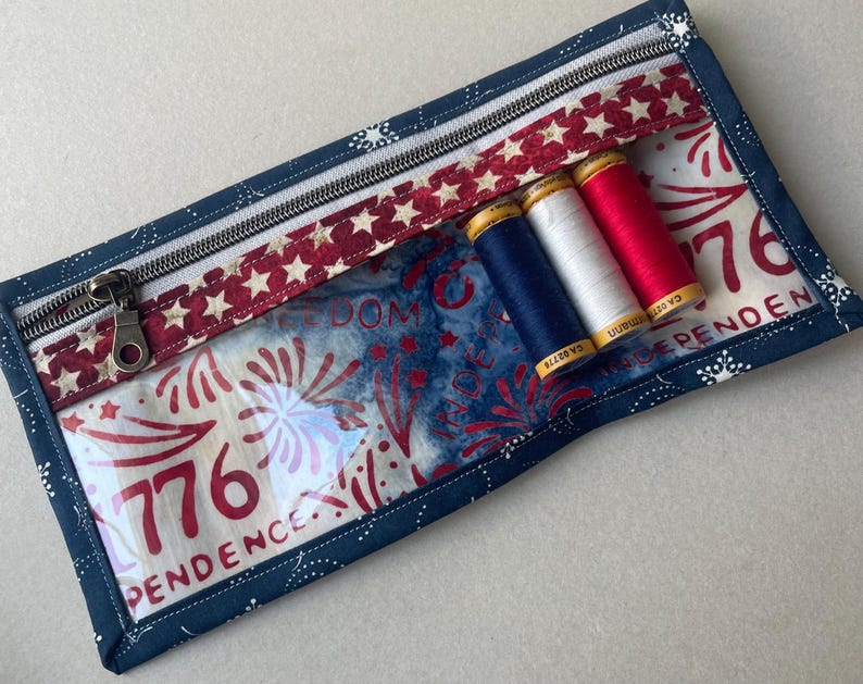 May include: A rectangular, patriotic-themed zippered pouch with a clear front panel. The pouch features a navy blue border, a red and white star-patterned accent, and a zipper. Inside are three spools of thread in red, white, and blue.