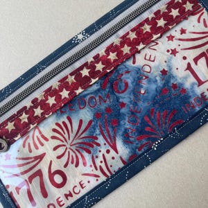 May include: Rectangular zippered pouch with a patriotic theme. The pouch features a red, white, and blue color scheme with star and firework designs. The text "1776 INDEPENDENCE" is visible on the front. The pouch is trimmed with a dark blue fabric.