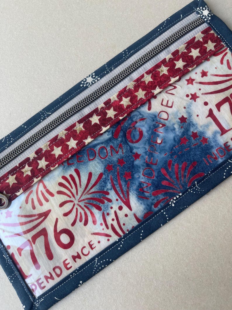 May include: Rectangular zippered pouch with a patriotic theme. The pouch features a red, white, and blue color scheme with star and firework designs. The text "1776 INDEPENDENCE" is visible on the front. The pouch is trimmed with a dark blue fabric.