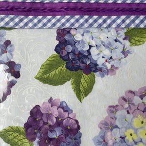 May include: Close-up of a fabric with a floral pattern featuring purple and lavender hydrangeas with green leaves. The top edge has a purple zipper and a blue and white gingham pattern.