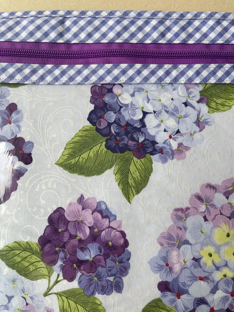 May include: Close-up of a fabric with a floral pattern featuring purple and lavender hydrangeas with green leaves. The top edge has a purple zipper and a blue and white gingham pattern.