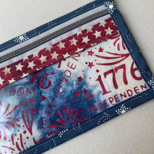 May include: A rectangular fabric item with a zipper, featuring a patriotic design. The design includes red stars on a white background, blue tie-dye, and the number "1776" in red. The edges are trimmed with a dark blue fabric.