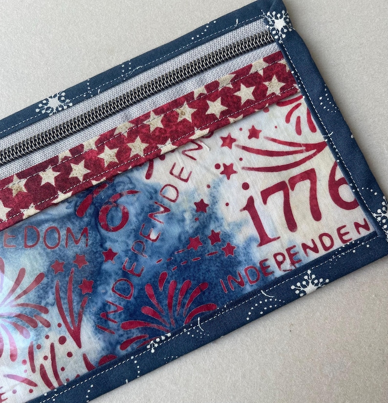 May include: A rectangular fabric item with a zipper, featuring a patriotic design. The design includes red stars on a white background, blue tie-dye, and the number "1776" in red. The edges are trimmed with a dark blue fabric.