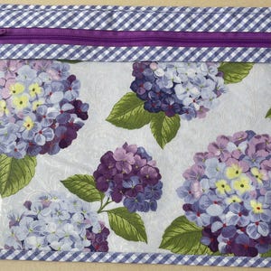 May include: A clear zippered pouch with a floral hydrangea design. The flowers are in shades of purple, lavender, and yellow, with green leaves, set against a white background. The pouch is trimmed with a blue and white gingham pattern and a purple zipper.
