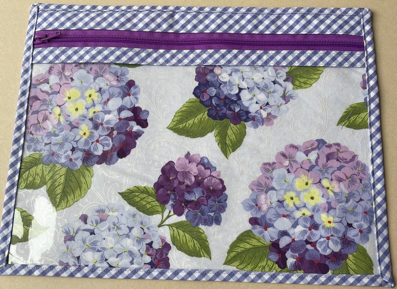 May include: A clear zippered pouch with a floral hydrangea design. The flowers are in shades of purple, lavender, and yellow, with green leaves, set against a white background. The pouch is trimmed with a blue and white gingham pattern and a purple zipper.