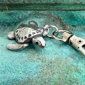 May include: A silver-colored turtle keychain with a lobster clasp. The turtle has detailed markings on its shell and flippers. The keychain is attached to a silver-colored metal clasp.