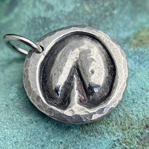 May include: A silver charm in the shape of a horse's hoof print. The charm has a hammered texture and a loop for attaching to a necklace or bracelet.