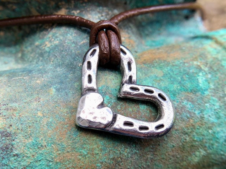 May include: A silver heart-shaped pendant necklace with a horseshoe design, hanging from a brown leather cord.