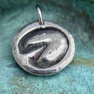 May include: A silver pendant with a textured, hammered finish. The pendant features a raised, three-dimensional design of a horse's hoof print.