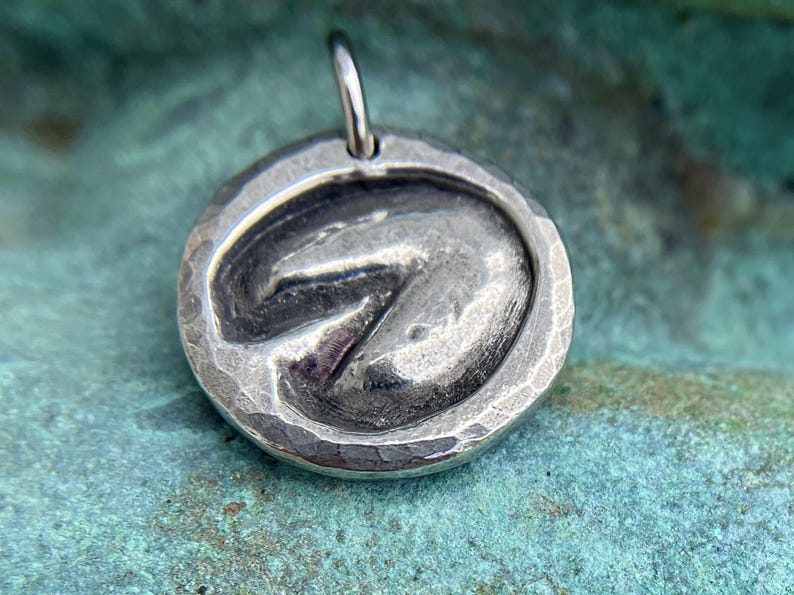 May include: A silver pendant with a textured, hammered finish. The pendant features a raised, three-dimensional design of a horse's hoof print.